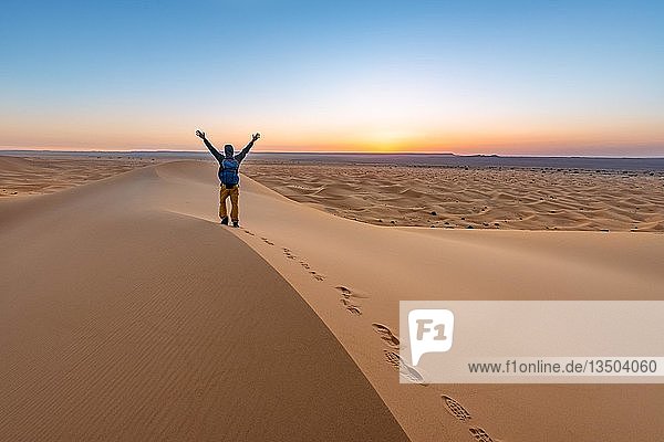 Junger Mann streckt seine Arme in die Luft  Sanddüne bei Sonnenaufgang  Erg Chebbi  Merzouga  Sahara  Marokko  Afrika
