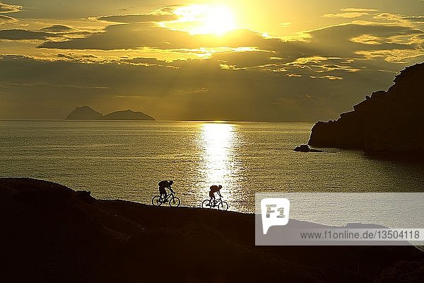 Two mountain bikers cycling on a rocky coast  back light at sunset  Red Beach  Matala  Crete  Greece  Europe