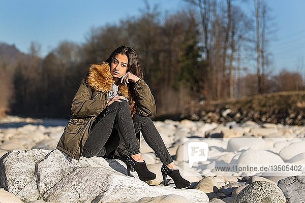 Young woman with long brunette hair sits on stone in dry river in winter  fashion  look into the camera