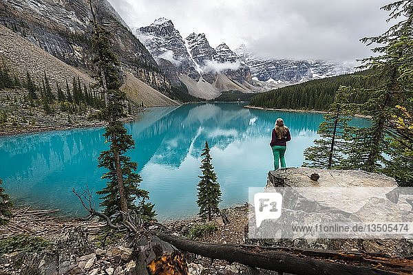 Young woman standing in front of a lake looking into mountain scenery  clouds hanging between mountain peaks  reflection in turquoise lake  Moraine Lake  Valley of the Ten Peaks  Rocky Mountains  Banff National Park  Province of Alberta  Canada  North America