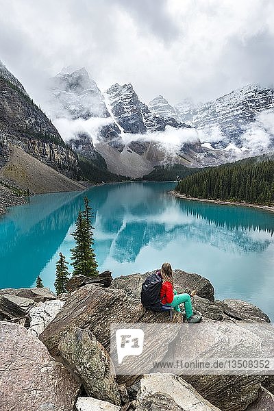 Junge Frau sitzt vor einem See und schaut in die Berglandschaft  Wolken hängen zwischen Berggipfeln  Spiegelung im türkisfarbenen See  Moraine Lake  Valley of the Ten Peaks  Rocky Mountains  Banff National Park  Provinz Alberta  Kanada  Nordamerika