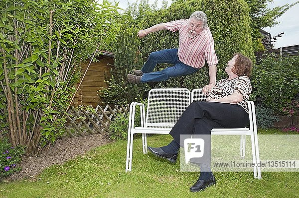 Ein älterer Bürger springt vor Freude über eine Bank  auf der seine Frau sitzt