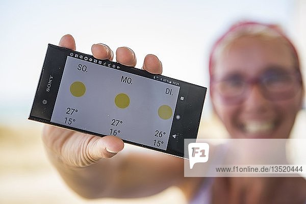 Lachende Frau am Strand freut sich über sonniges Wetter im Wetterbericht auf ihrem Smartphone  Normandie  Frankreich  Europa