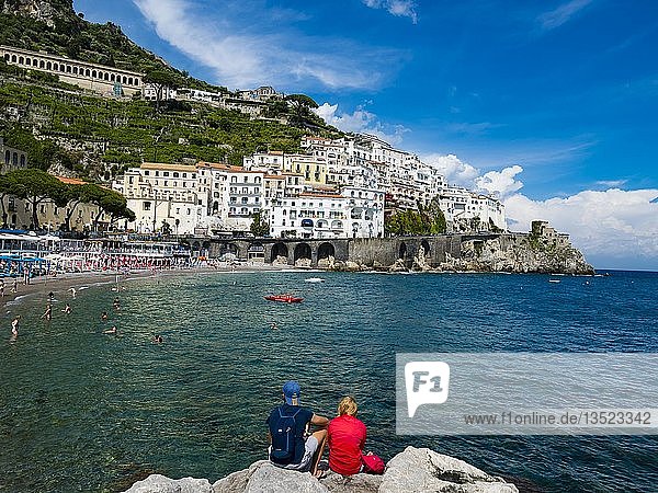 Couple sits on rock and looks at old town and beach of Amalfi  peninsula of Sorrento  Amalfi coast  Campania  Italy  Europe
