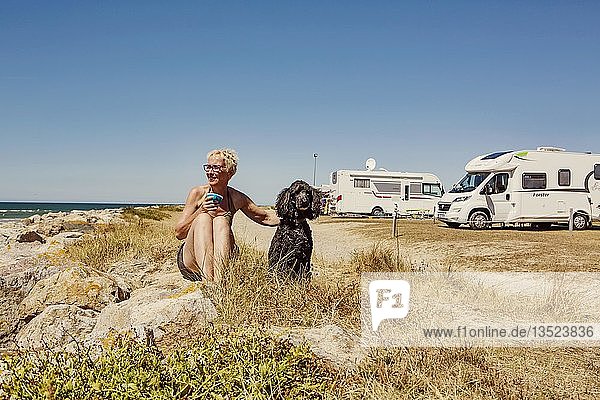 Frau mit sitzt mit ihrem Hund  Königspudel  im Gras auf einem Wohnmobilstellplatz  Strand am Meer  Portbail  Normandie  Frankreich  Europa