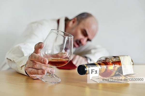 Alkoholsüchtiger  der ein Cognacglas hält und mit einer Flasche Alkohol an einem Tisch sitzt