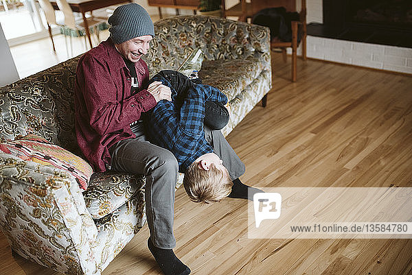 Verspielter Vater hält seinen Sohn kopfüber auf dem Wohnzimmersofa