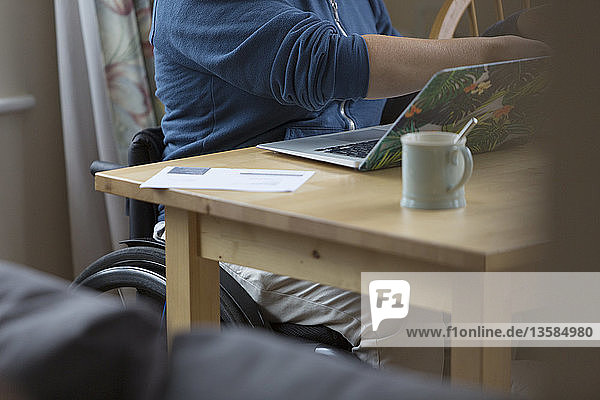 Junge Frau im Rollstuhl mit Laptop am Esstisch