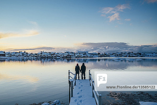 Couple holding hands at the edge of snowy dock overlooking waterfront village  Reine  Lofoten Islands  Norway