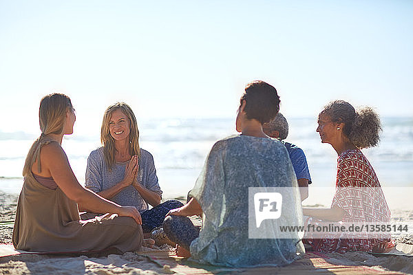 Glückliche Freunde unterhalten sich im Kreis am sonnigen Strand während eines Yoga-Retreats