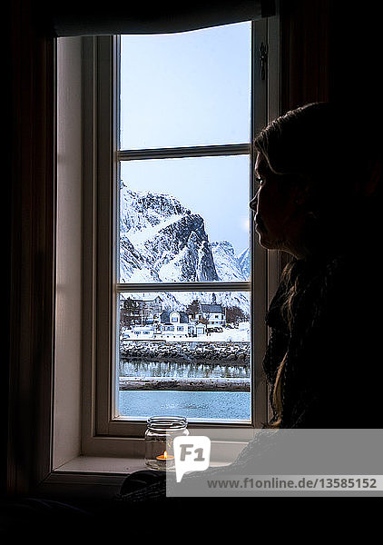 Nachdenkliche Frau schaut aus dem Fenster auf einen verschneiten Berg  Reine  Lofoten  Norwegen
