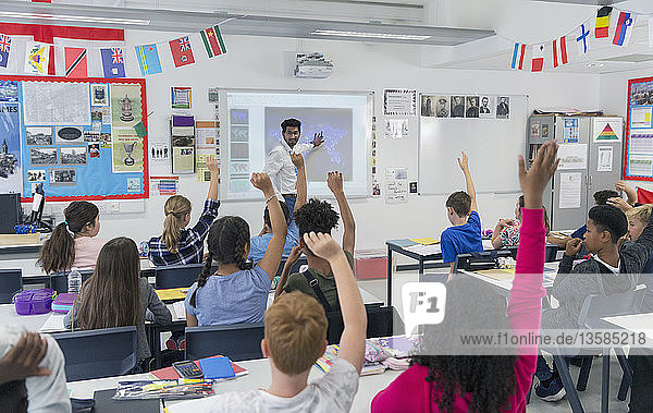 Männlicher Lehrer  der eine Unterrichtsstunde an der Projektionsfläche im Klassenzimmer leitet  während die Schüler die Hände heben