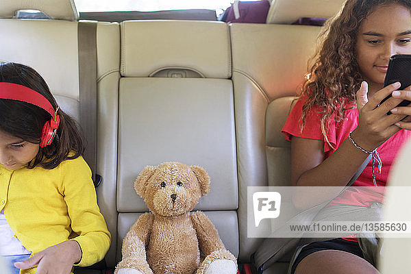 Schwestern mit Teddybär  die ein Smartphone und ein digitales Tablet benutzen und auf dem Rücksitz eines Autos sitzen