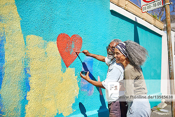 Happy senior couple painting heart-shape mural on sunny wall