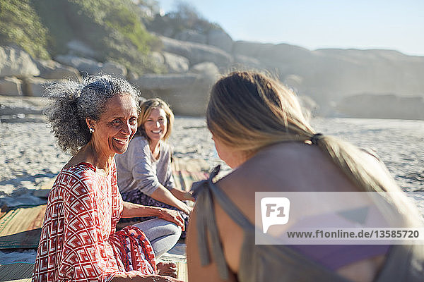 Lächelnde Freundinnen unterhalten sich am sonnigen Strand während eines Yoga-Retreats