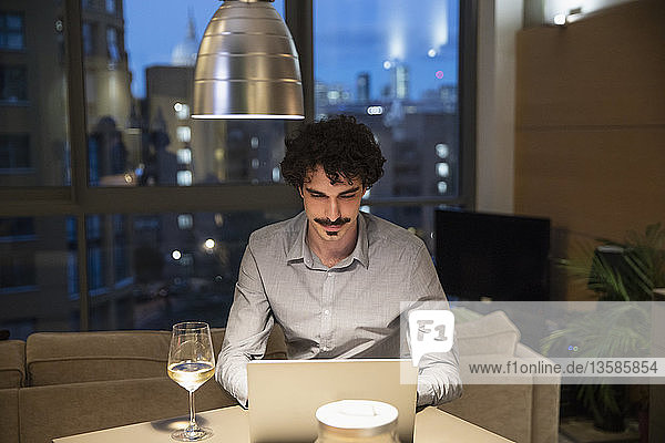 Mann benutzt Laptop und trinkt Weißwein in der Küche einer städtischen Wohnung bei Nacht