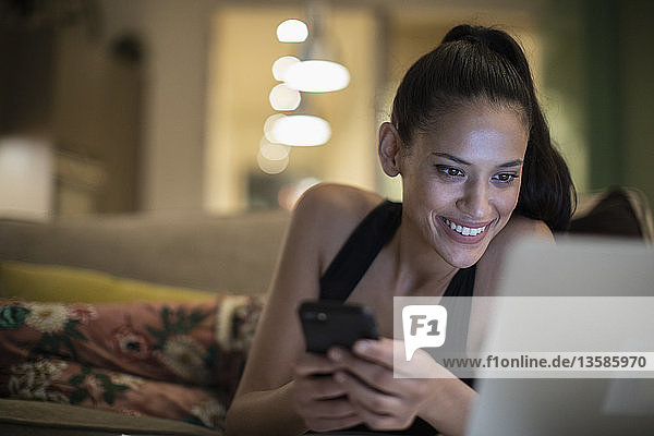 Smiling woman in pajamas using smart phone and laptop on sofa
