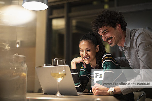 Glückliches Paar benutzt einen Laptop und trinkt Weißwein in einer Wohnung bei Nacht