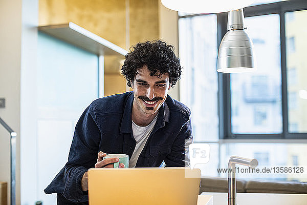Lächelnder Mann trinkt Kaffee und arbeitet am Laptop in der Wohnküche