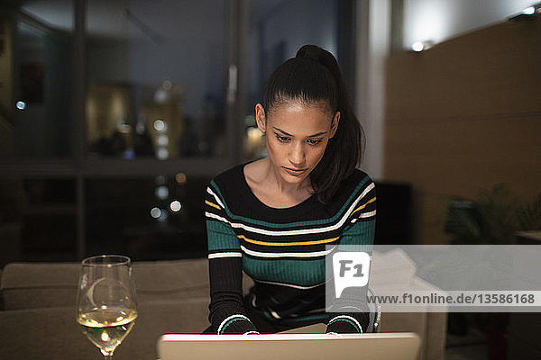 Konzentrierte Frau trinkt Weißwein und arbeitet abends zu Hause am Laptop