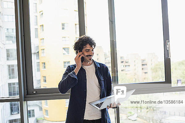 Mann spricht mit Smartphone und benutzt Laptop am Fenster einer städtischen Wohnung