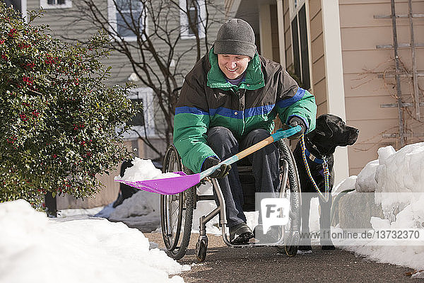 Frau mit Multipler Sklerose schaufelt Schnee im Rollstuhl mit einem Diensthund