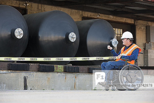 Gebäudetechniker im Rollstuhl beim Ziehen eines Absperrbandes vor einem Chemielagertank