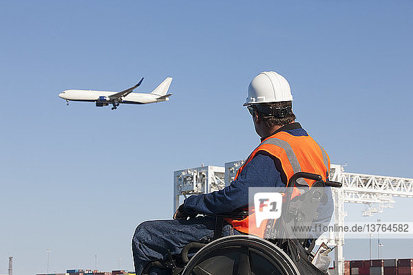Verkehrsingenieur im Rollstuhl beobachtet ein Flugzeug  das über Schiffscontainer und Kräne im Hafen fliegt