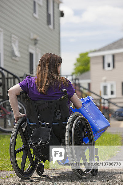 Frau mit Spina Bifida im Rollstuhl beim Abholen einer Recycling-Tonne an der Straße