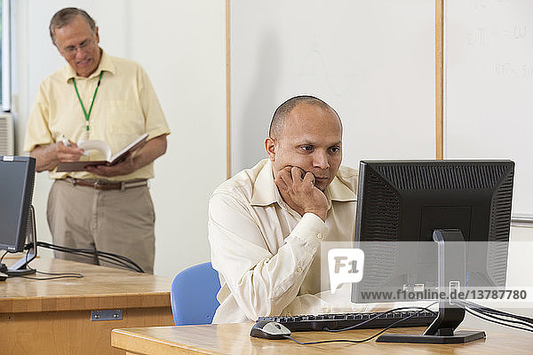 Engineering student studying computer display in a classroom with professor in background