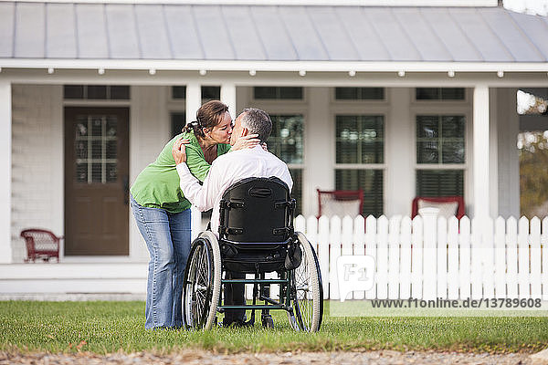 Pärchen küsst sich vor ihrem neuen Haus  während er im Rollstuhl sitzt