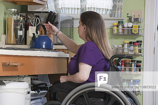 Frau mit Spina Bifida im Rollstuhl beim Aufstellen eines Wasserkochers in der barrierefreien Küche