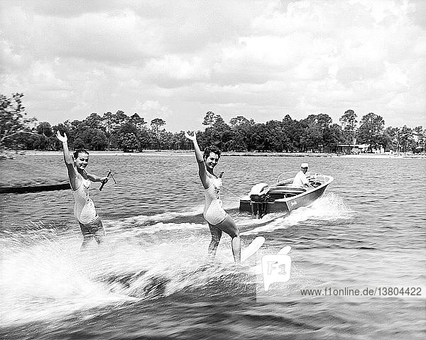 Vereinigte Staaten: ca. 1959 Zwei junge Frauen  die auf einem See Wasserski fahren  winken der Kamera zu  als sie vorbeifahren.