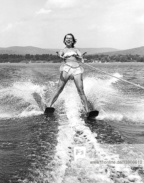 Vereinigte Staaten: um 1955 Eine junge Frau mit einem breiten Lächeln auf dem Gesicht  während sie dicht hinter dem Boot Wasserski fährt.