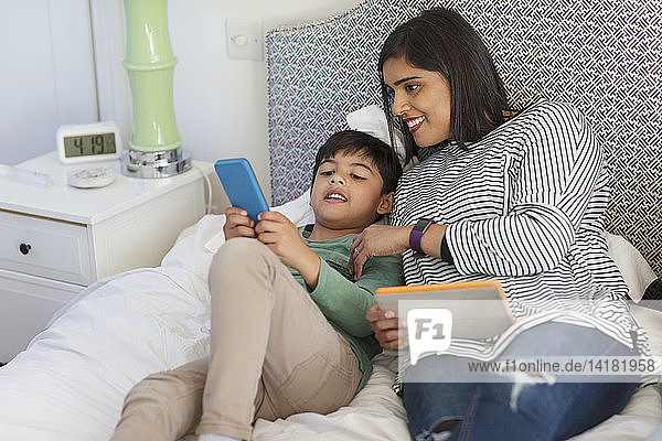 Mother and son using smart phone and digital tablet on bed