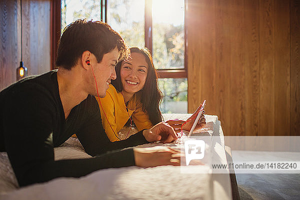 Lächelndes Paar mit Kopfhörern  das sich ein digitales Tablet im Bett teilt