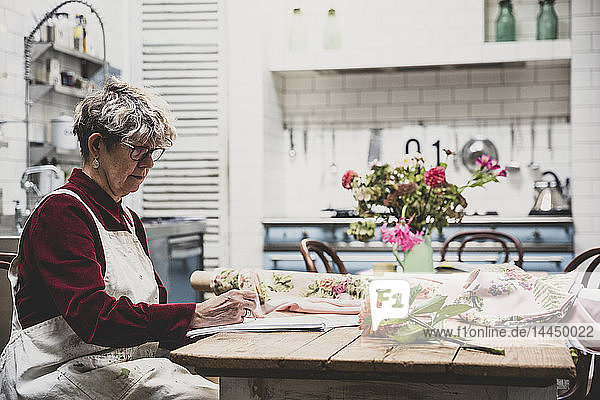 Ältere Frau mit Brille  rotem Kleid und weißer Schürze sitzt am Tisch und arbeitet an einer Bleistiftzeichnung der orangefarbenen Dahlie.