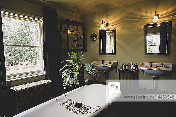 Innenansicht des Badezimmers mit schwarzen Spiegeln über zwei viktorianischen Waschtischen  Schiebefenster und Roll-Top-Badewanne mit Messing-Badewagen.
