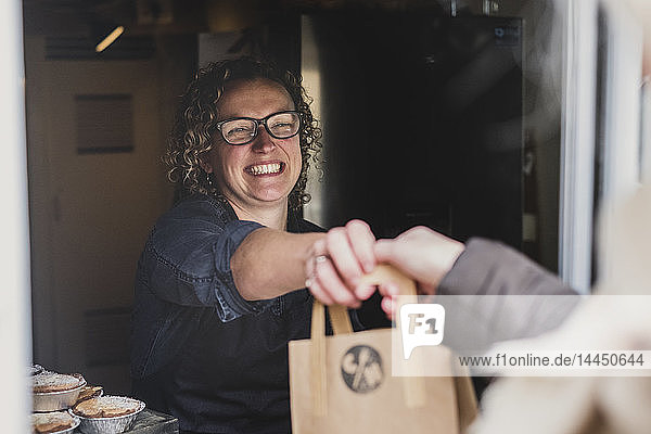 Lächelnde Frau mit Brille  die eine braune Einkaufstasche aus Papier durch das Fenster einer Bäckerei reicht.