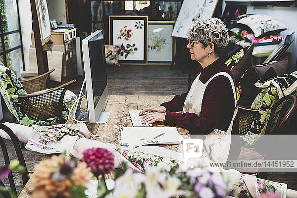 Ältere Frau mit Brille  rotem Kleid und weißer Schürze sitzt an einem Holztisch in einem Studio und arbeitet am Desktop-Computer.