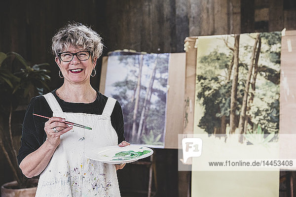 Ältere Frau mit Brille  schwarzem Oberteil und weißer Schürze steht im Atelier und arbeitet an der Bemalung von Bäumen im Wald.