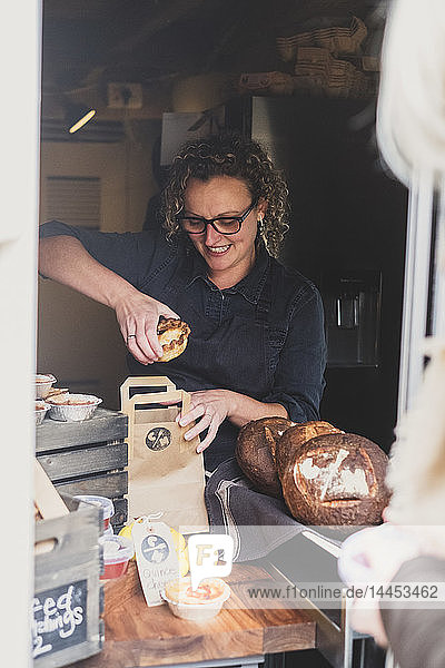 Lächelnde Frau mit Brille legt frisch gebackenen Kuchen in eine Einkaufstasche aus braunem Papier.