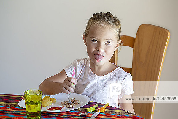 Hübsches kleines Mädchen am Tisch vor ihrem Teller und versucht  ihre Zunge herauszuziehen.