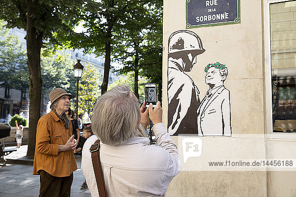 Rückenansicht einer Person beim Fotografieren eines Straßenkunstwerks  Reproduktion des Fotos von Cohn Bendit und CRS-Helm Christian Le Padellec  aufgenommen vor der Sorbonne im Mai 68.
