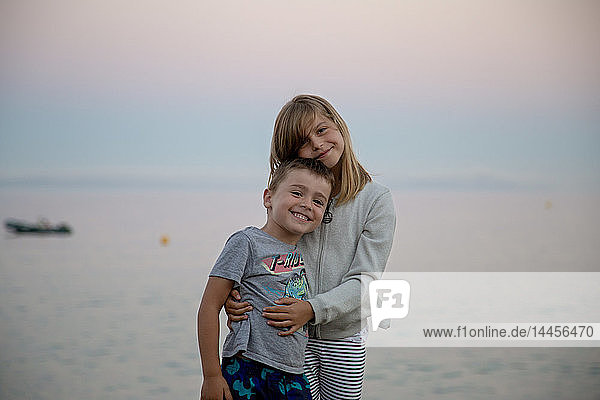 A little girl with her brother in the arms posing tenderly in front of the sea at nightfall.