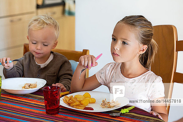 A little girl eating potatoes and chicken with her little brother sitting at the table.