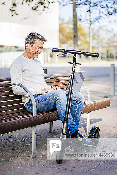 Casual businessman with kick scooter  sitting on a bench  working relaxed in the city