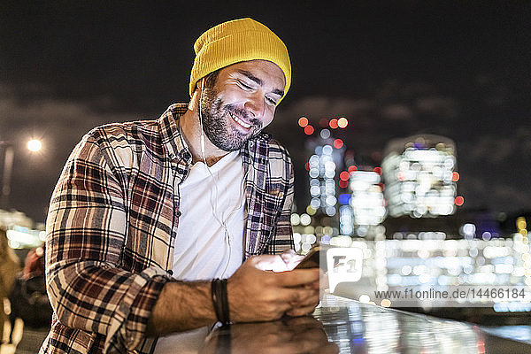 Großbritannien  London  lächelnder Mann lehnt an einem Geländer und schaut auf sein Telefon mit Stadtlichtern im Hintergrund