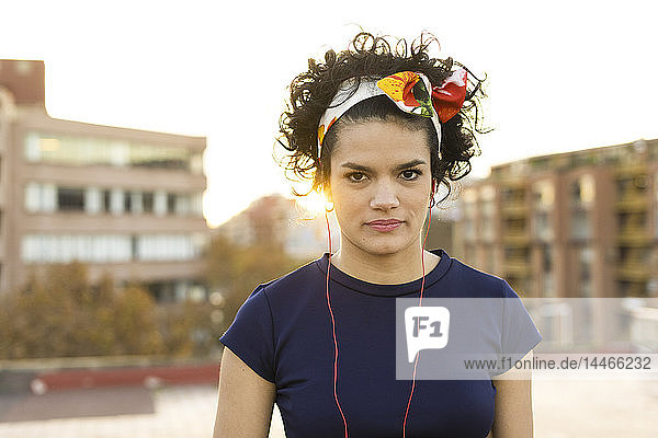Porträt einer jungen Frau mit Haarreif und Kopfhörer in der Stadt bei Sonnenuntergang
