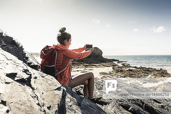 Junge Frau sitzt an einem felsigen Strand und benutzt ein Smartphone